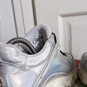 Op de afbeelding: Een paar zilveren, iriserende platform sneakers met witte zolen. De binnenkant van de schoen heeft een wit label met de tekst "Buffalo" in zwart.