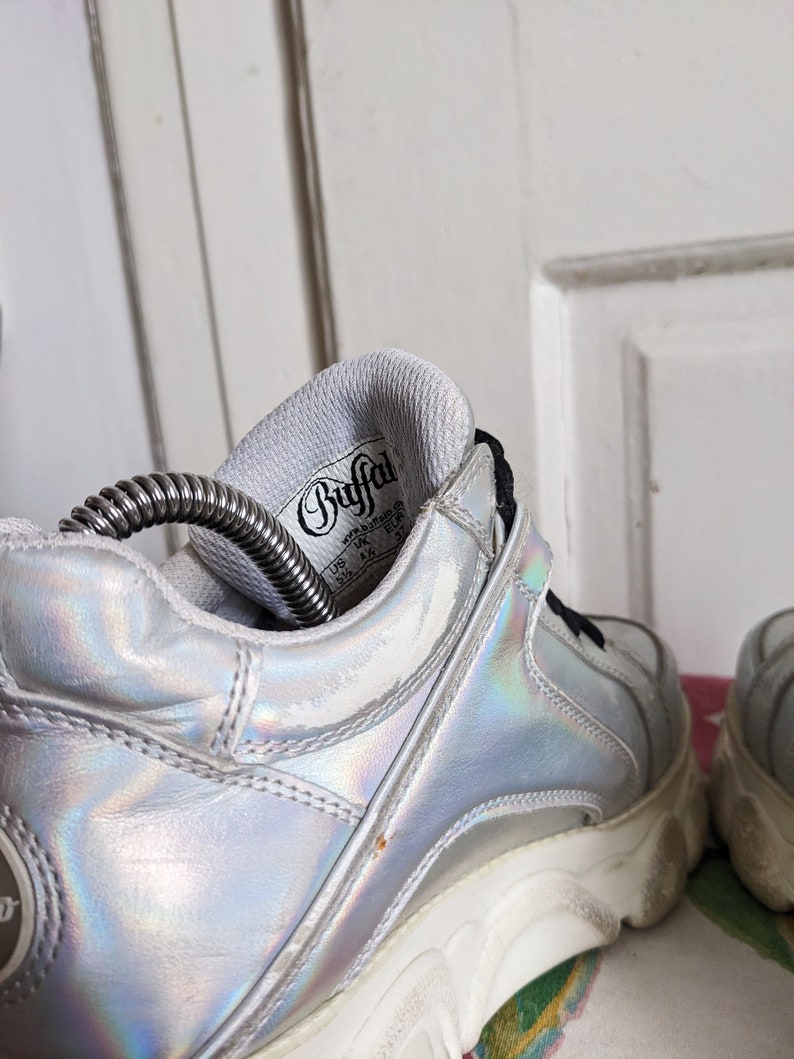 Op de afbeelding: Een paar zilveren, iriserende platform sneakers met witte zolen. De binnenkant van de schoen heeft een wit label met de tekst "Buffalo" in zwart.