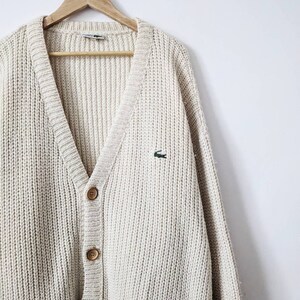 May include: A cream-colored knitted cardigan with a V-neck, button closure, and a small green crocodile logo on the left chest.