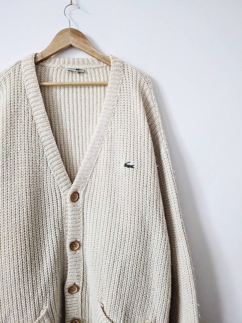 May include: A cream-colored knitted cardigan with a V-neck, button closure, and a small green crocodile logo on the left chest.