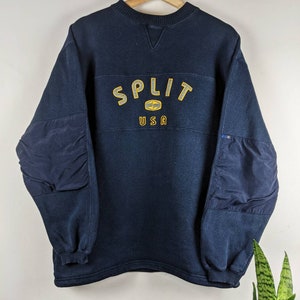 以下が含まれることがあります： 前面に「SPLIT USA」と刺繍された黄色いロゴが付いたネイビーのトレーナー。トレーナーにはネイビーの肘当てが付いています。