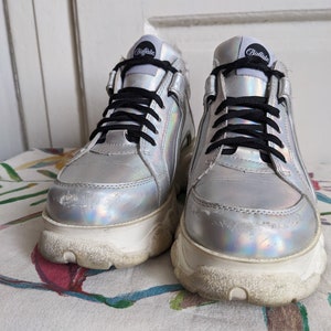 Op de afbeelding: Een paar zilveren platform sneakers met zwarte veters en het woord "Buffalo" gedrukt op de zijkant.