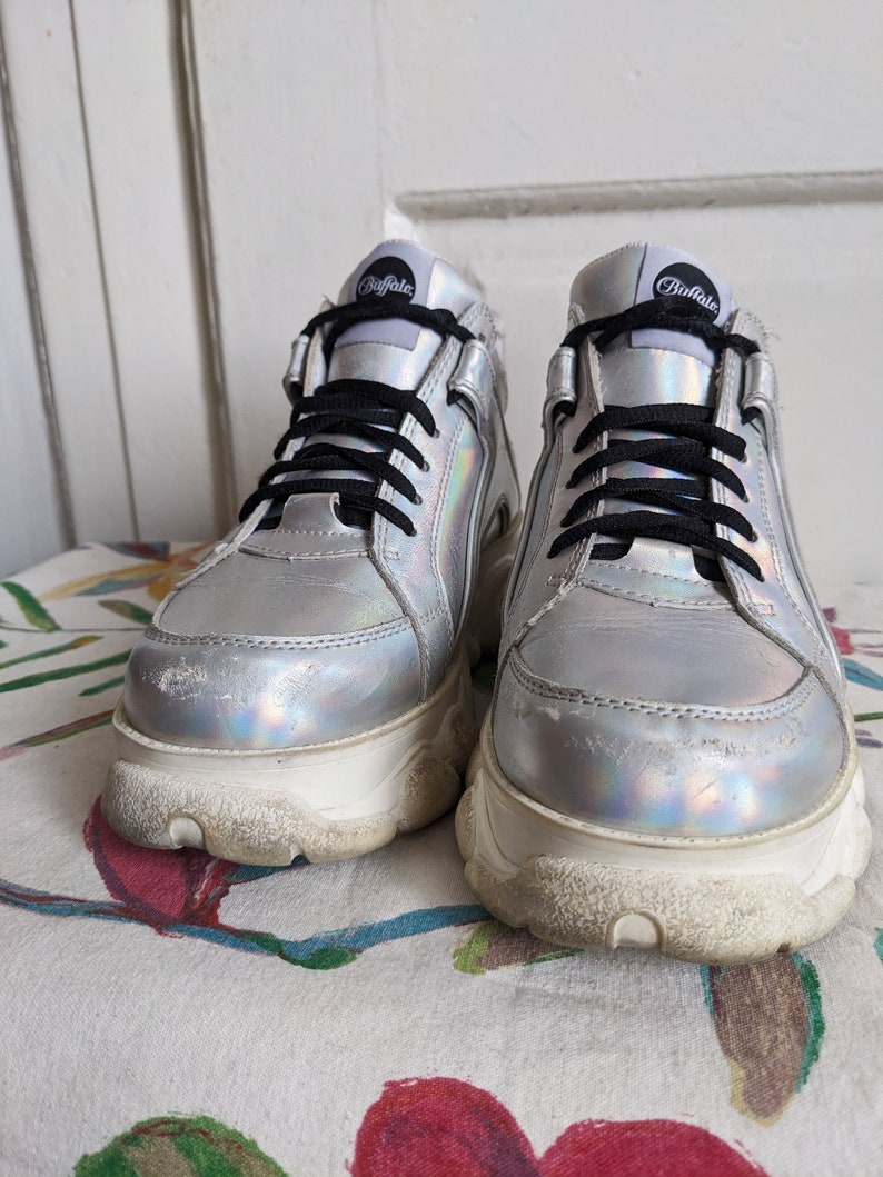 Op de afbeelding: Een paar zilveren platform sneakers met zwarte veters en het woord "Buffalo" gedrukt op de zijkant.