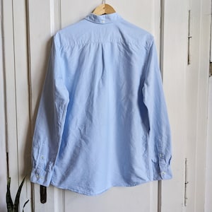 May include: A light blue long-sleeve button-down shirt hanging on a wooden hanger. The shirt has a classic collar, buttoned cuffs, and a curved hem. The back of the shirt is visible, showing the yoke and pleats.