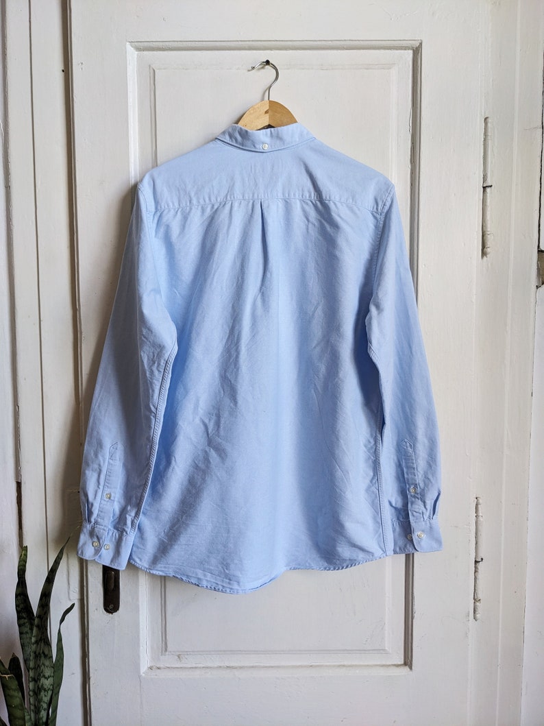 May include: A light blue long-sleeve button-down shirt hanging on a wooden hanger. The shirt has a classic collar, buttoned cuffs, and a curved hem. The back of the shirt is visible, showing the yoke and pleats.