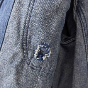 May include: Close-up of a blue denim jacket with a small hole in the elbow area. The hole is surrounded by stitching.