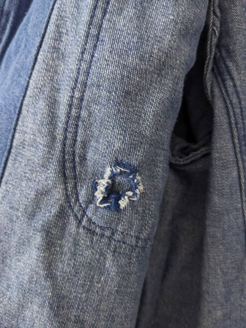 May include: Close-up of a blue denim jacket with a small hole in the elbow area. The hole is surrounded by stitching.