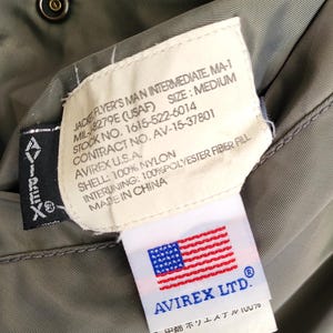 Puede incluir: Primer plano de la etiqueta interior de una chaqueta verde militar. La etiqueta dice "JACKET FLYER'S MAN INTERMEDIATE, MA-1" e incluye detalles sobre las especificaciones de la chaqueta, incluido el fabricante, la talla y los materiales.