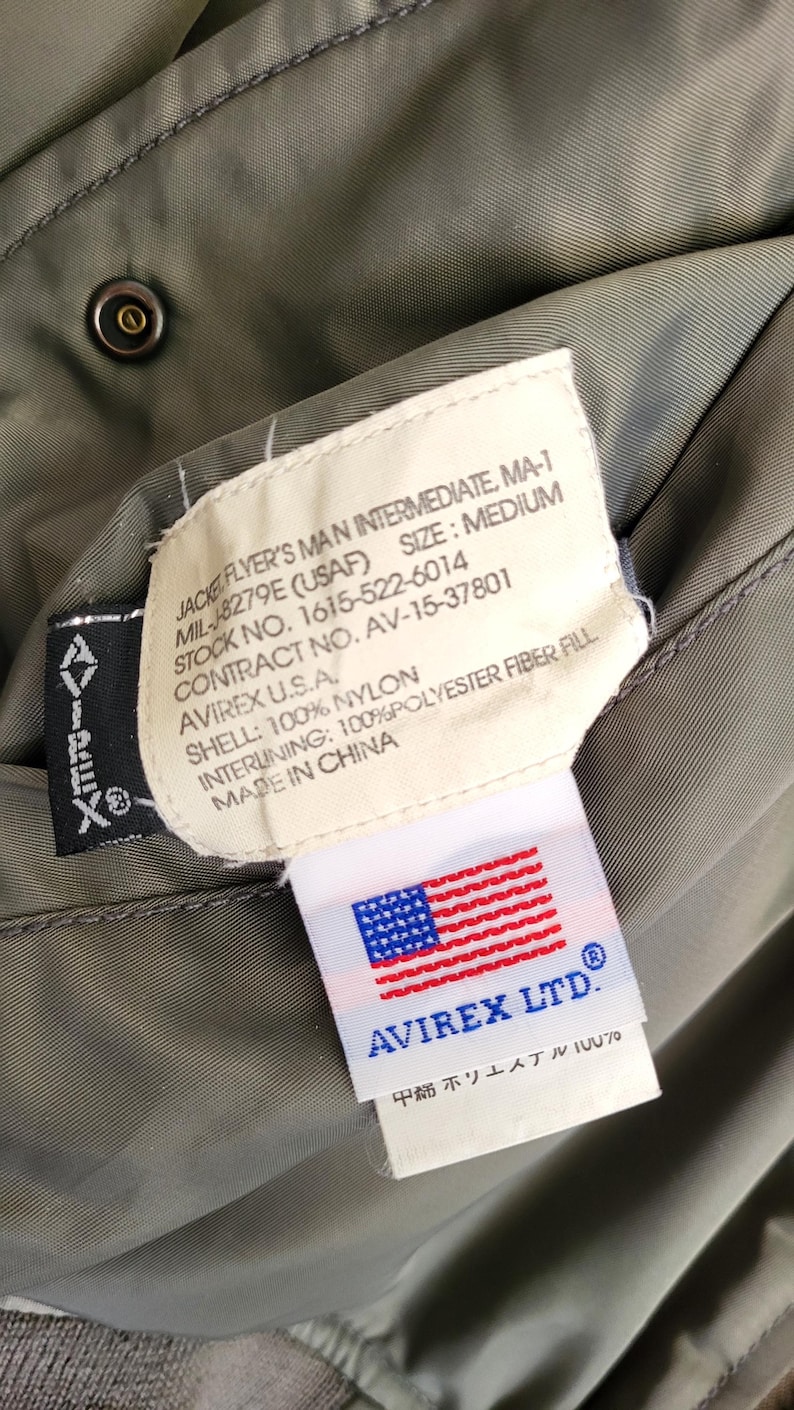 Puede incluir: Primer plano de la etiqueta interior de una chaqueta verde militar. La etiqueta dice "JACKET FLYER'S MAN INTERMEDIATE, MA-1" e incluye detalles sobre las especificaciones de la chaqueta, incluido el fabricante, la talla y los materiales.
