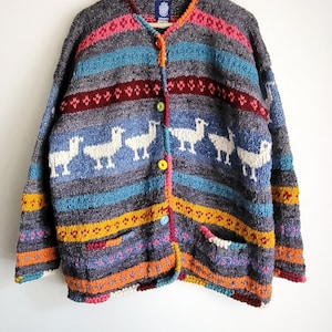 May include: A multi-coloured, button-up cardigan with a llama pattern. The sweater has horizontal stripes in grey, blue, pink, and orange. It features two pockets and is suspended from a wooden hanger.