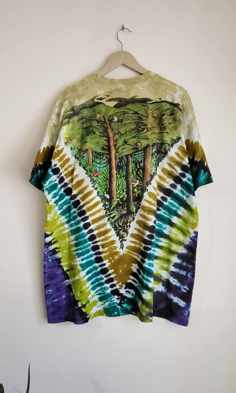 Puede incluir: Una camiseta te&ntilde;ida con un dise&ntilde;o de bosque en la espalda. La camiseta presenta un patr&oacute;n te&ntilde;ido en forma de V en tonos morado, verde, amarillo y marr&oacute;n. La escena del bosque representa &aacute;rboles y follaje.