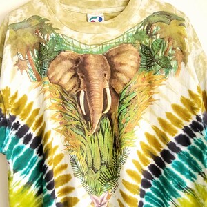 Puede incluir: Una camiseta te&ntilde;ida con un dise&ntilde;o de elefante rodeado de exuberante follaje verde y palmeras. La camiseta tiene una base blanca con un colorido patr&oacute;n te&ntilde;ido en tonos de amarillo, verde y morado.