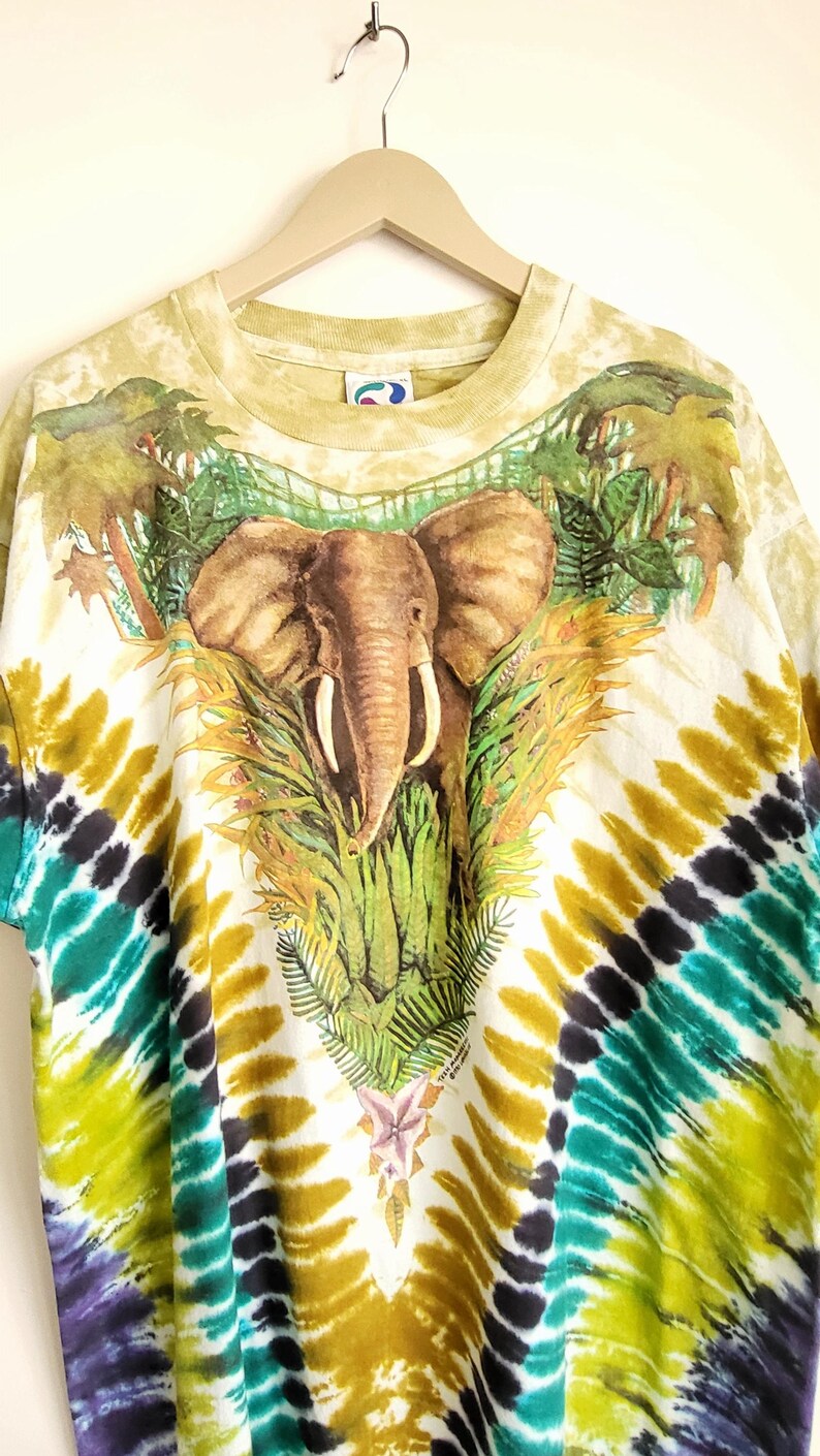 Puede incluir: Una camiseta te&ntilde;ida con un dise&ntilde;o de elefante rodeado de exuberante follaje verde y palmeras. La camiseta tiene una base blanca con un colorido patr&oacute;n te&ntilde;ido en tonos de amarillo, verde y morado.