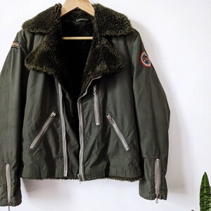 May include: A green jacket with a faux fur collar and zippered pockets. The jacket has two patches on the front, one with the word "AVIATORS" and the other with a circular design.