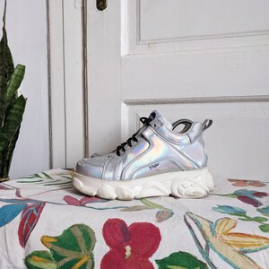 Op de afbeelding: Zilveren, iriserende platform sneakers met een dikke witte zool. De merknaam "Vans" is zichtbaar aan de zijkant van de schoen.