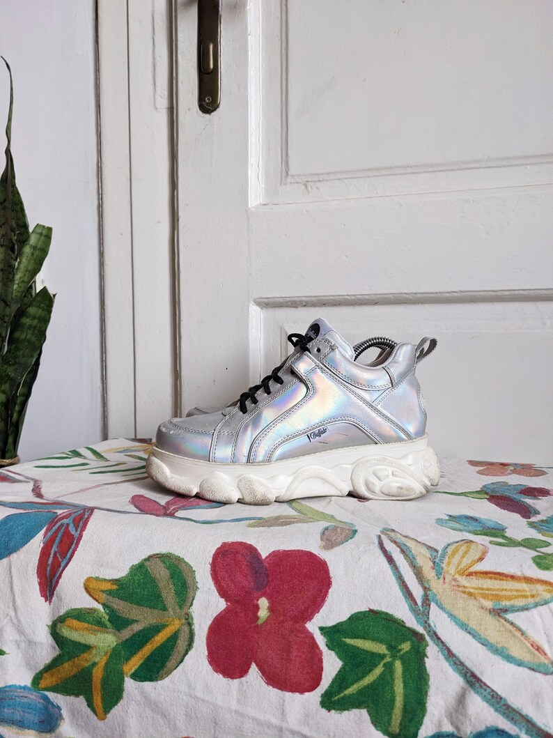 Op de afbeelding: Zilveren, iriserende platform sneakers met een dikke witte zool. De merknaam "Vans" is zichtbaar aan de zijkant van de schoen.