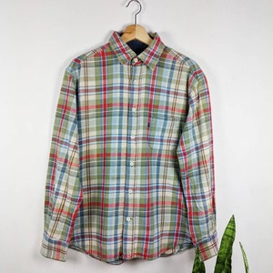 May include: A long-sleeved, button-down plaid shirt. The shirt has a pattern of red, green, blue, and cream checks. It is displayed on a wooden hanger against a white backdrop. The shirt is a classic style.