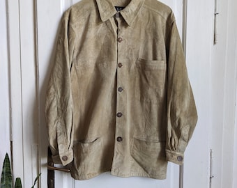 Pode incluir: Uma camisa de camurça marrom claro com fecho de botões e vários bolsos. A camisa está pendurada em um cabide de madeira contra uma porta branca.