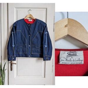 May include: A blue denim jacket with a red lining and blue stitching. The jacket has a button closure and a label that reads "Levi's Studio by Jean Charles de Castelbajac".