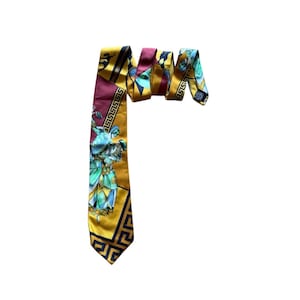 Puede incluir: Una corbata de seda con un patrón geométrico y ilustraciones de figuras con vestimenta clásica. Los colores son dorado, burdeos y verde azulado. La corbata mide aproximadamente 147 cm de largo.