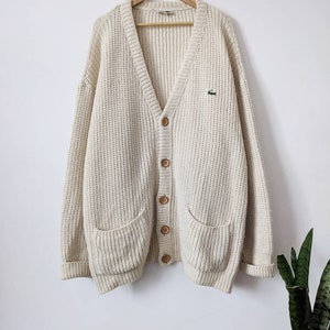 May include: A cream-colored knitted cardigan with a V-neck, button closure, and two patch pockets. The cardigan features a small green crocodile logo on the left chest.