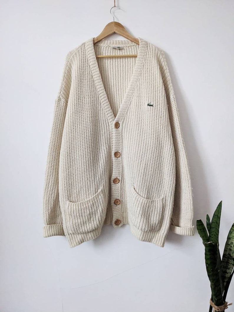 May include: A cream-colored knitted cardigan with a V-neck, button closure, and two patch pockets. The cardigan features a small green crocodile logo on the left chest.