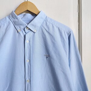May include: A light blue button-down shirt with a classic collar and long sleeves, hanging on a wooden hanger. The shirt features a small embroidered logo on the chest. The shirt is made of a smooth, woven fabric.