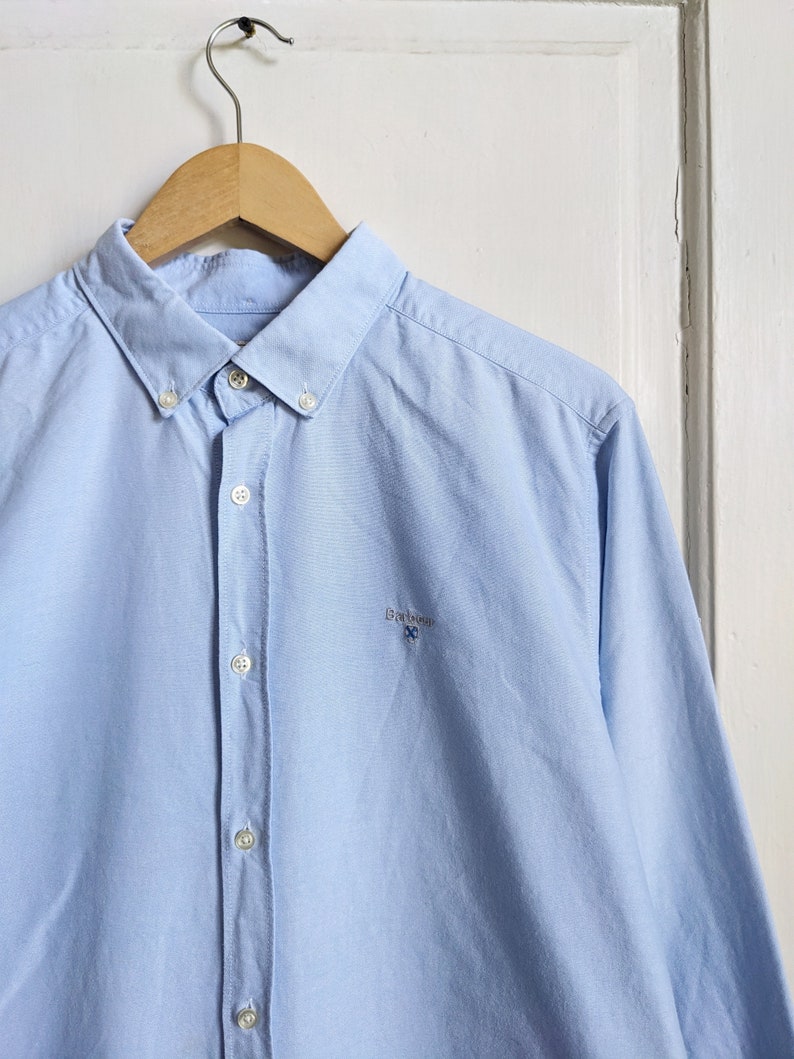 May include: A light blue button-down shirt with a classic collar and long sleeves, hanging on a wooden hanger. The shirt features a small embroidered logo on the chest. The shirt is made of a smooth, woven fabric.