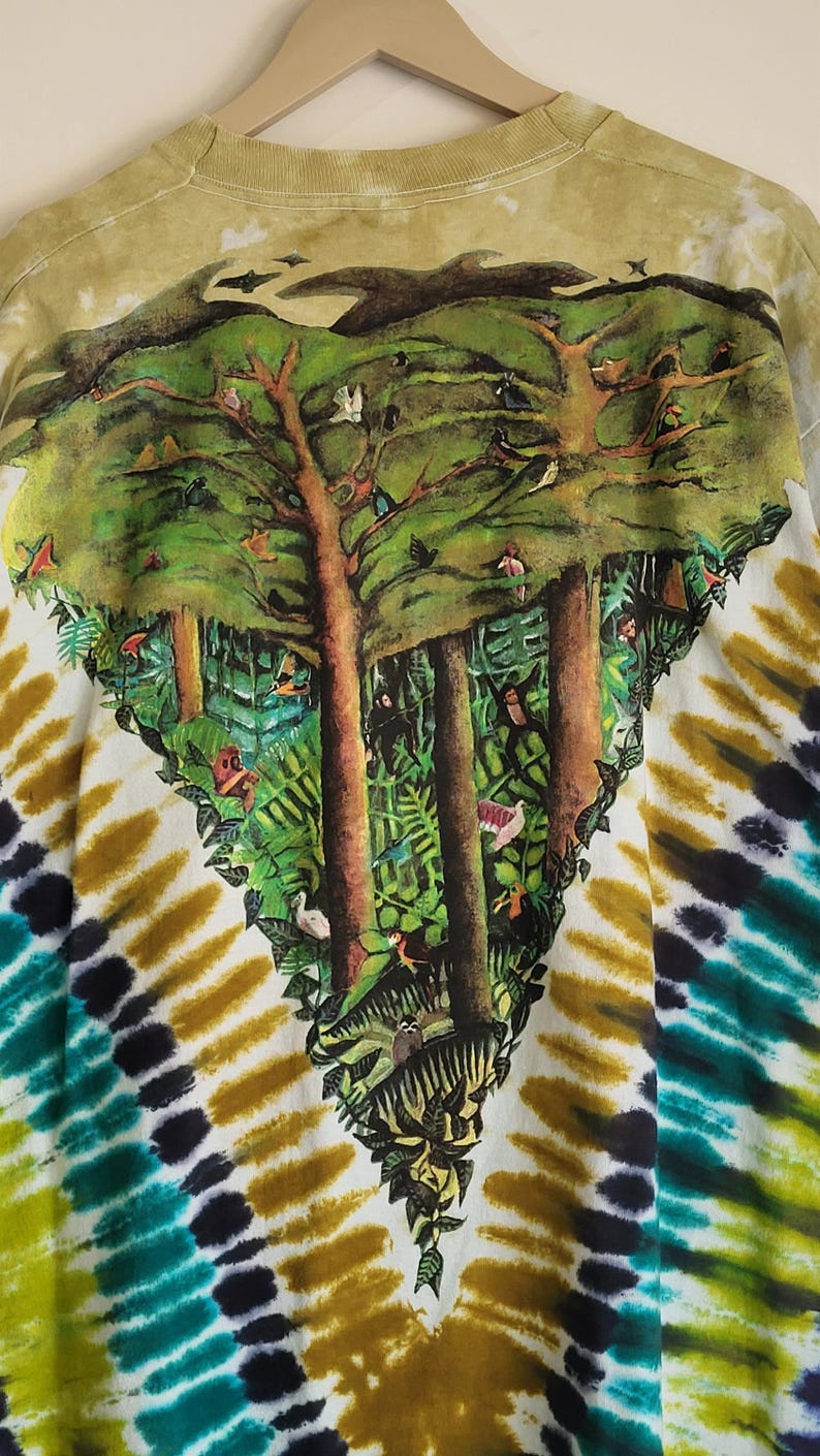 Puede incluir: Una camiseta te&ntilde;ida con un dise&ntilde;o de la naturaleza. El dise&ntilde;o presenta una escena de bosque con &aacute;rboles, p&aacute;jaros y vegetaci&oacute;n exuberante. El patr&oacute;n tie-dye de la camisa incluye tonos de verde, amarillo y azul.