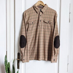 Pode incluir: Uma camisa de manga comprida xadrez em marrom, preto e branco. A camisa tem dois bolsos com aba na frente e patches de camurça marrom nos cotovelos.
