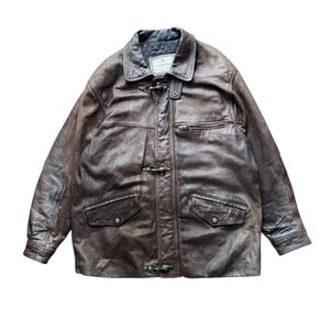 Può includere: Giacca in pelle marrone con collo, due tasche frontali e chiusure a toggle. La giacca ha un aspetto invecchiato e un'etichetta sul colletto. La giacca è in stile vintage.