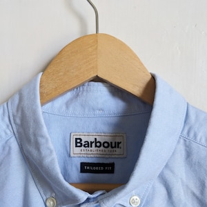 May include: A light blue button-down shirt with a classic collar, hanging on a wooden hanger. The shirt features the "Barbour" label and the words "Tailored Fit". The shirt is made of a light, possibly cotton, fabric.
