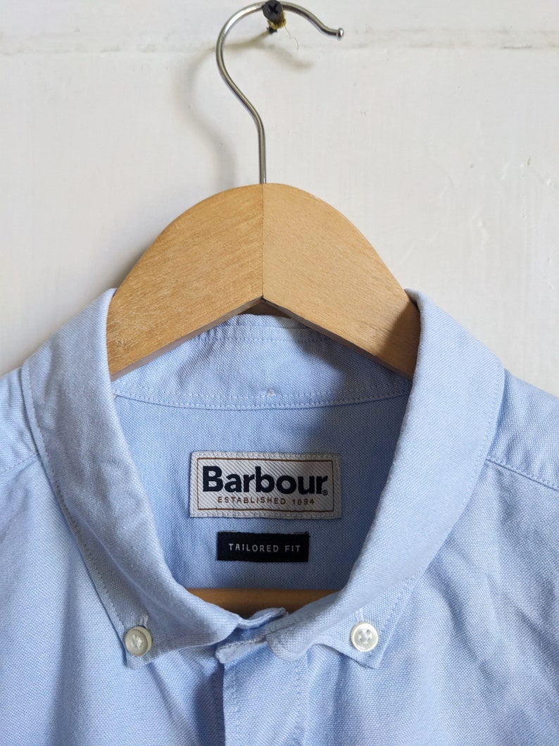 May include: A light blue button-down shirt with a classic collar, hanging on a wooden hanger. The shirt features the "Barbour" label and the words "Tailored Fit". The shirt is made of a light, possibly cotton, fabric.