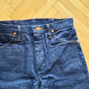 Op de afbeelding: Close-up van een donkerblauwe denim jeans. De jeans heeft koperkleurige klinknagels en een knoopsluiting. De stiksels zijn in een contrasterende oranje kleur. De jeans is een klassieke stijl.
