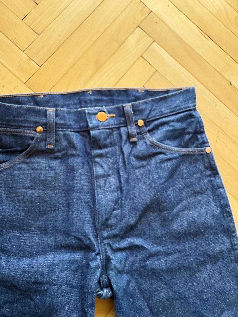 Op de afbeelding: Close-up van een donkerblauwe denim jeans. De jeans heeft koperkleurige klinknagels en een knoopsluiting. De stiksels zijn in een contrasterende oranje kleur. De jeans is een klassieke stijl.