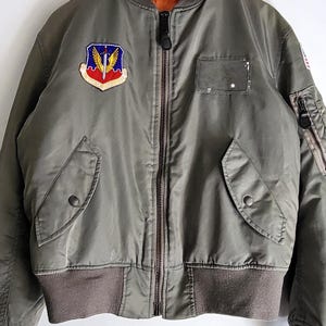 Puede incluir: Chaqueta bomber verde oliva con cuello, pu&ntilde;os y cintura de canal&eacute; marr&oacute;n. Cuenta con un parche con un escudo y alas, cierre de cremallera y dos bolsillos delanteros. La chaqueta est&aacute; colgada de un gancho de metal.