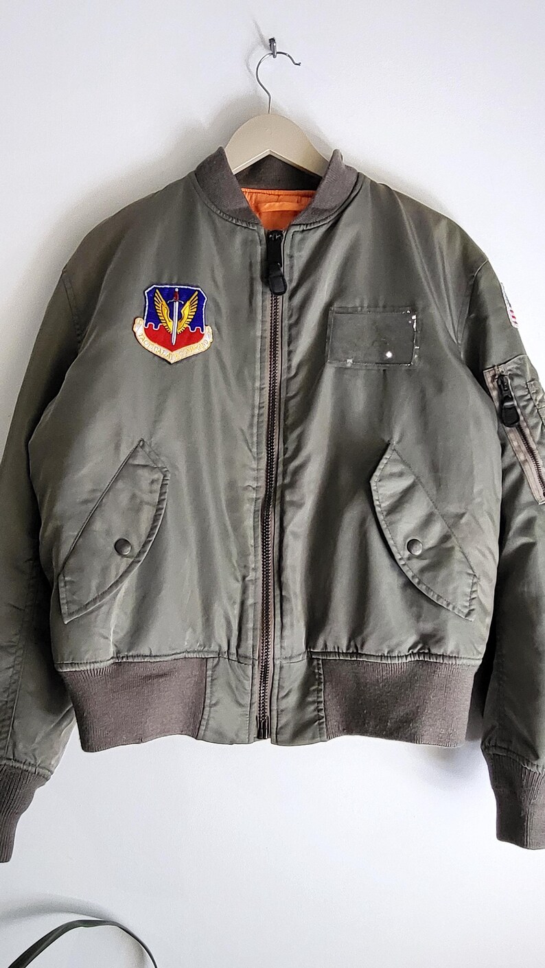 Puede incluir: Chaqueta bomber verde oliva con cuello, pu&ntilde;os y cintura de canal&eacute; marr&oacute;n. Cuenta con un parche con un escudo y alas, cierre de cremallera y dos bolsillos delanteros. La chaqueta est&aacute; colgada de un gancho de metal.