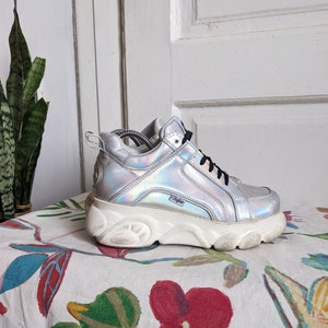 Op de afbeelding: Zilveren platform sneakers met een dikke witte zool. De schoenen hebben een holografische afwerking en een zwart logo aan de zijkant.
