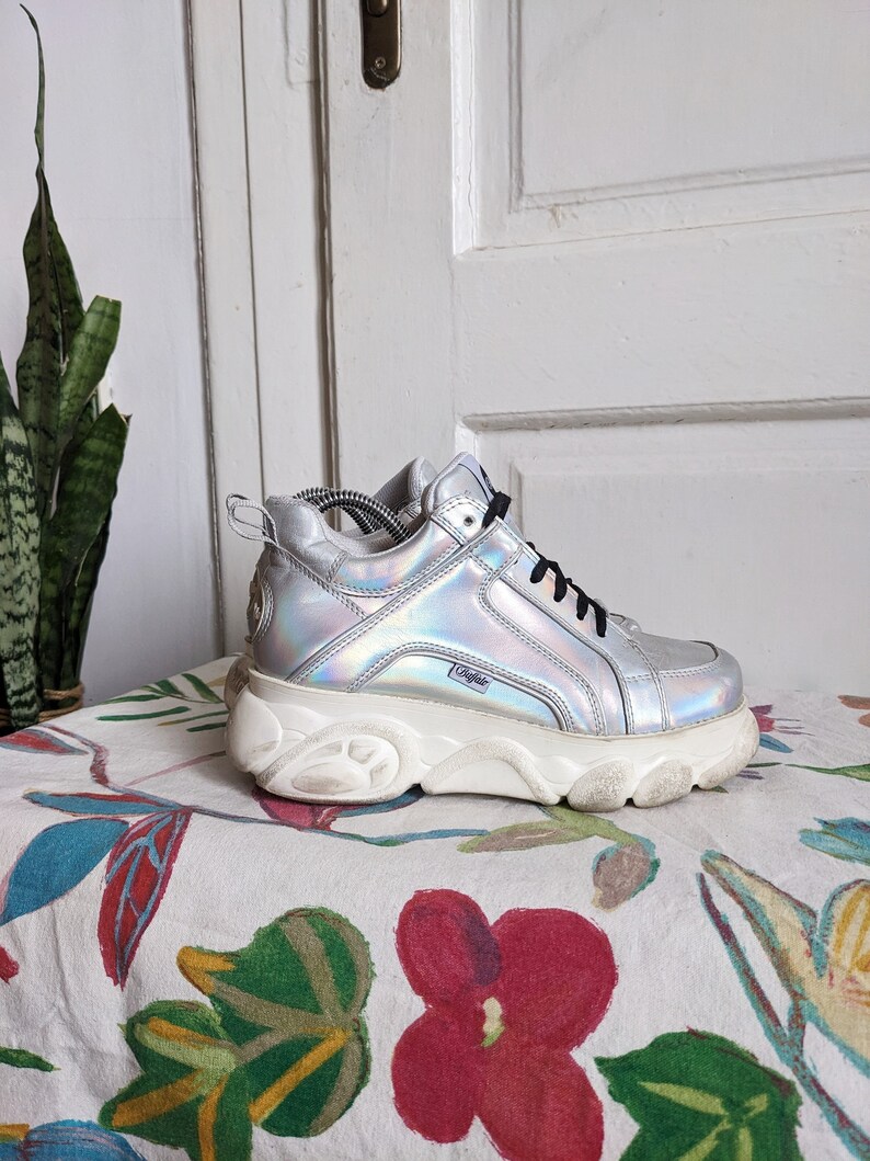 Op de afbeelding: Zilveren platform sneakers met een dikke witte zool. De schoenen hebben een holografische afwerking en een zwart logo aan de zijkant.