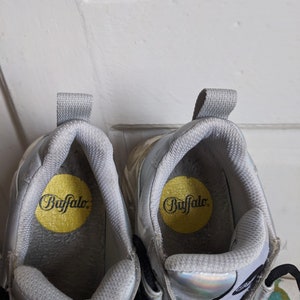 Op de afbeelding: Een paar zilveren platform sneakers met zwarte veters en een gele binnenzool met de tekst "Buffalo".