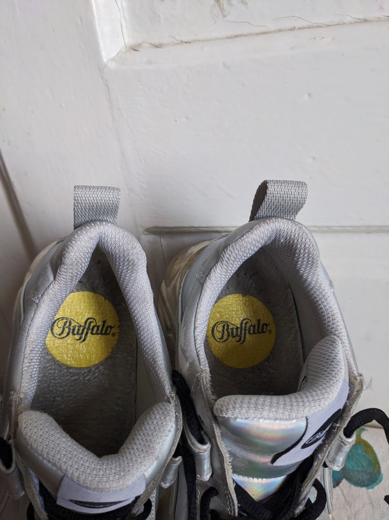Op de afbeelding: Een paar zilveren platform sneakers met zwarte veters en een gele binnenzool met de tekst "Buffalo".