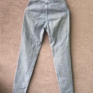 K&ouml;nnte beinhalten: Hellblaue Acid-Wash-Jeans mit hoher Taille und geradem Bein. Auf der R&uuml;ckseite befinden sich zwei Taschen und ein schwarzer Lederaufn&auml;her mit silbernem Emblem. Die Jeans h&auml;ngen an einem Metallb&uuml;gel.