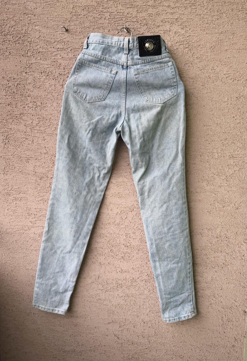 K&ouml;nnte beinhalten: Hellblaue Acid-Wash-Jeans mit hoher Taille und geradem Bein. Auf der R&uuml;ckseite befinden sich zwei Taschen und ein schwarzer Lederaufn&auml;her mit silbernem Emblem. Die Jeans h&auml;ngen an einem Metallb&uuml;gel.