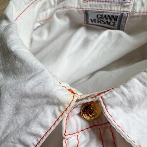 May include: A white button-down shirt featuring bright orange contrast stitching along the collar and placket. The shirt includes gold-toned metal buttons and interior labels reading Gianni Versace and Made in Italy, visible at the neckline.