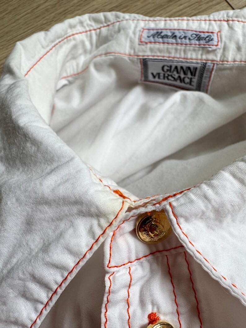 May include: A white button-down shirt featuring bright orange contrast stitching along the collar and placket. The shirt includes gold-toned metal buttons and interior labels reading Gianni Versace and Made in Italy, visible at the neckline.
