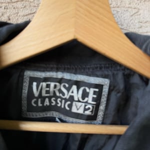 Puede incluir: Primer plano de una chaqueta negra con la etiqueta Versace Classic V2 visible. La etiqueta es rectangular con letras blancas. La chaqueta est&aacute; colgada en una percha de madera.