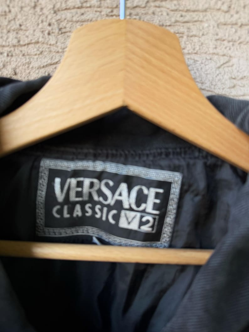 Puede incluir: Primer plano de una chaqueta negra con la etiqueta Versace Classic V2 visible. La etiqueta es rectangular con letras blancas. La chaqueta est&aacute; colgada en una percha de madera.