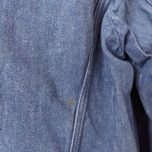 May include: Close-up of a blue denim fabric with a seam running diagonally across the image. The fabric has a slightly textured surface and appears to be worn.