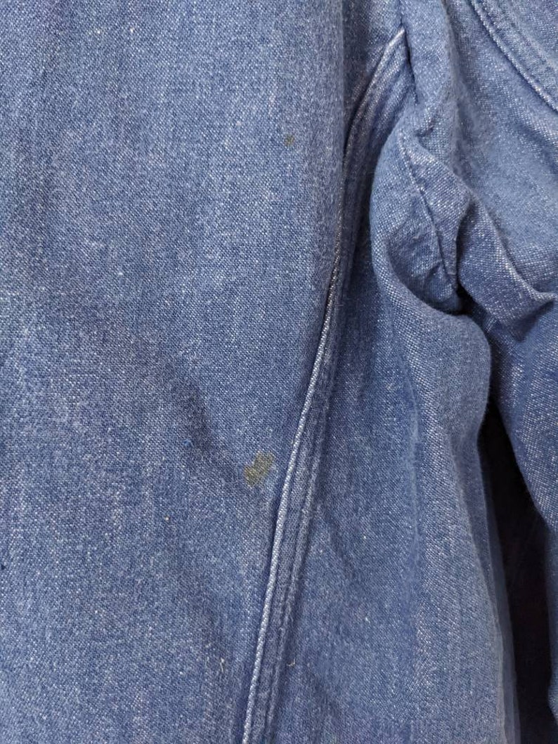 May include: Close-up of a blue denim fabric with a seam running diagonally across the image. The fabric has a slightly textured surface and appears to be worn.