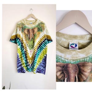 Puede incluir: Una camiseta te&ntilde;ida con un dise&ntilde;o de elefante. La camiseta tiene un patr&oacute;n en forma de V en tonos azules, verdes y marrones. El gr&aacute;fico del elefante est&aacute; centrado en la parte delantera, con una etiqueta Liquid Blue visible. La camiseta est&aacute; colgada en una percha de madera.